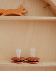 A wooden shelf holds a small orange fox figure on the top and two FRIGG Daisy Silicone Pacifiers (2-Pack) made from medical-grade silicone with clear nipples below, all set against a light wood background for a cozy, comforting baby scene.