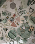 A flat lay of mushie pastel green and pink baby items, including plates, cups, bibs, teethers, pacifier clips, utensils, soft fabric pieces like the Organic Cotton Muslin Burp Cloth 2-Pack by mushie—all arranged on a white surface.