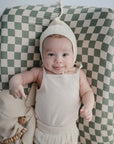 A baby in a cream knit bonnet and sleeveless top smiles while lying on the mushie Extra Soft Muslin Changing Pad Cover, featuring a green and white checkered pattern. A beige cloth and wooden toy rest nearby.