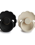 The FRIGG Daisy Natural Rubber Pacifier 2-Pack includes one black and one cream daisy-shaped pacifier, each with a latex nipple, round handle, scalloped edges, and FRIGG branding at the base.