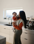 A woman stands in a sunlit modern kitchen holding a smiling baby in a striped sweater, who grasps the Mushie Trainer Sippy Cup with easy-grip handles.