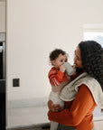 A woman with long curly hair smiles at a toddler in the kitchen as sunlight streams in. The toddler drinks from the Mushie Trainer Sippy Cup, which features easy-grip handles.