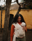 A smiling woman with curly hair carries a baby in a cream wrap carrier outdoors. The baby's outfit features the mushie Silicone Pacifier Clip | Eva, offering stylish and safe pacifier accessory near a tree and yellow wall.