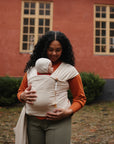 A woman with curly hair stands outside wearing an orange top and green pants, holding a baby in a beige wrap. The baby wears a hood and chews on the mushie Silicone Pacifier Clip | Eva. A red building with large windows is behind them.