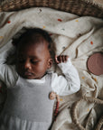 A baby sleeps peacefully in a woven basket lined with the mushie Organic Cotton Muslin Swaddle Blanket, while a round teether toy rests nearby on the cream patterned blanket.