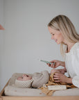 A smiling woman holds a phone while changing her baby—dressed in a knit bonnet and sweater—on the mushie Extra Soft Muslin Changing Pad Cover. Colorful fabric pennants hang in the background as the baby looks up at her.