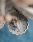 Close-up of the FRIGG Andersen Fairytale Silicone Pacifier, a decorative circular white pacifier with heart-shaped cutouts, set against a soft blue background. Sold as a 2-pack from the FRIGG brand.