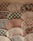 The mushie Organic Cotton Muslin Burp Cloth 2-Pack features rounded burp cloths in checks, florals, paisley, and solid muted beige, pink, green, and cream tones arranged in a scalloped pattern.