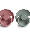 The FRIGG Daisy Silicone Pacifier 2-Pack includes two scalloped-edge pacifiers in dusty pink and muted green, each with a circular handle and embossed FRIGG logo, made from medical grade silicone for baby comfort.