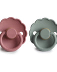 The FRIGG Daisy Natural Rubber Pacifier 2-Pack includes scalloped-edge daisy-shaped pacifiers in dusty pink and muted green, each with natural rubber nipples, circular handles, and the FRIGG brand embossed on the bottom.