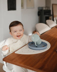 A smiling baby in a bright, modern room sits at a wooden table wearing a bib, holding the Mushie Trainer Sippy Cup upside down over a plate with crumbs scattered on the table.