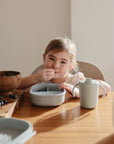 A young child sits at a wooden table, eating from a divided bowl. Nearby, a Mushie Trainer Sippy Cup with easy-grip handles and a bowl of blueberries sit on the table in the sunlight.