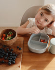 A smiling young girl at a wooden table with a grey silicone divided plate of strawberries, a Mushie Trainer Sippy Cup with easy-grip handles, a wooden bowl of strawberries, and fresh blueberries on a cutting board.