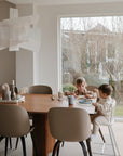 Two young children sit at a wooden dining table in a bright, modern room. One child drinks from the Mushie Trainer Sippy Cup with easy-grip handles and non-toxic material while the other eats amid minimalist decor.