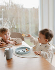 Two young children sit at a table by a large window, eating snacks. One offers food on a fork to the other. Plates, food, and a Mushie Trainer Sippy Cup with easy-grip handles are on the table.