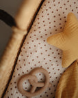 A close-up of a woven basket with a white sheet featuring small brown floral patterns, a yellow star cushion, and the mushie Pretzel Teether in beige silicone.