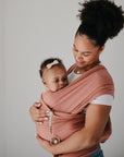A woman smiles, holding her baby close in a pink wrap. The relaxed baby wears a headband and a mushie Silicone Pacifier Clip | Eva as they stand against a plain white background.