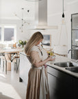 In a sunlit modern kitchen, a woman in a light-patterned dress and baby wrap uses the mushie Silicone Pacifier Clip | Eva for her infant while preparing something at the sink.