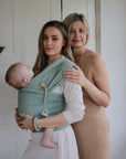 A young woman holds a sleeping baby in a green wrap, showcasing the mushie Silicone Pacifier Clip | Eva, while an older woman stands beside them. Both smile gently at the camera in a bright, airy room.