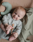 A smiling baby with light hair lies on the mushie Moon Lovey Blanket, holding a wooden bead toy and touching their foot. The baby wears a light gray outfit, with a green cloth nearby.