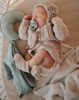 A baby sleeps in a crib, cuddling the mushie Moon Lovey Blanket with a pacifier, while wearing a knitted sweater, bonnet, and booties. Nearby are a soft blanket and cushion.