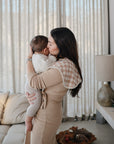 A woman in a beige dress holds and kisses a baby in a cream sweater, with mushie Organic Cotton Muslin Burp Cloth 2-Pack nearby, in a cozy living room with a sofa, lamp, and light curtains in the background.