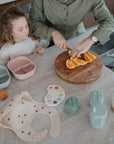 A woman slices oranges on a round wooden cutting board as a young girl watches. On the counter are divided plates, cookies, and a Mushie Trainer Sippy Cup with easy-grip handles, suggesting preparation for a child's meal.