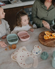A woman slices oranges on a round wooden board as two young girls watch at the counter. Nearby are divided snack containers, cookies, a bib, toy cups like the Mushie Trainer Sippy Cup with easy-grip handles, and a paint palette.