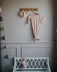 A soft pink baby gown and mushie Ribbed Baby Bonnet hang on wooden hooks above a white bench in a gently lit nursery, with a pastel garland draped along the wall.