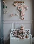 A baby wearing a knitted bonnet and cute outfit sits on a white bench in a pastel nursery, surrounded by soft toys, a mug, the mushie Moon Lovey Blanket made from organic cotton, and decorative bunting hanging on the wall above.