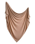 A large, beige Stretchy Swaddle by mushie, made from silky breathable fabric, is draped in elegant, swagged folds against a white background.
