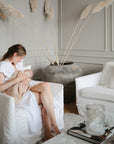 A woman in a white dress breastfeeds her baby wrapped in a mushie Organic Cotton Muslin Swaddle Blanket, sitting on a white armchair in a modern living room with neutral decor, pampas grass, and a nearby white sofa.