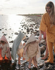 A woman in mustard holds hands with three small children in hooded towels, one displaying the mushie Silicone Pacifier Clip | Eva, as they walk barefoot on a rocky shoreline under a cloudy sky.