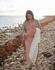 A pregnant woman in a long, flowing dress walks barefoot on a rocky beach, gently cradling her belly and holding a patterned blanket. The mushie Silicone Pacifier Clip | Eva peeks out from her bag as calm water stretches behind her.