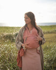 A woman stands in a grassy field near the water, cradling her baby in a pink wrap carrier. Beside the baby's legs rests a FRIGG Daisy Night Silicone Pacifier from FRIGG, adding sweetness to the peaceful, sunlit scene.