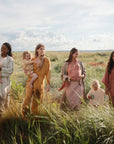 Four women and three children stroll through tall grass under a blue sky, with one child holding the mushie Silicone Pacifier Clip | Eva—an earth-toned, stylish pacifier accessory that complements their natural setting and clothing.