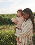 A woman and young child stand in a sunny grassy field, both in neutral-toned outfits. The child holds the mushie Silicone Pacifier Clip | Eva, with partly cloudy skies above.