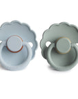 The FRIGG Daisy Natural Rubber Pacifier 2-Pack features light blue and green scallop-edged pacifiers with round handles, each designed for gentle comfort with a natural rubber latex nipple.