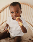 A toddler sits in a wicker chair, wearing a white shirt and patterned shorts, holding the mushie Flower Training Toothbrush (yellow) in their mouth and a pacifier in the other hand, against a neutral background.