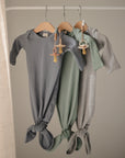 Three baby sleeper gowns in gray, green, and light gray hang on wooden hangers, each paired with the mushie Silicone Pacifier Clip | Eva. They are tied at the bottom and displayed on a simple rod in a softly lit room.