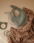 Two mushie Muslin Bibs—one light green, one beige—are displayed on leopard-print fabric next to two beaded teething rings and a green bear-shaped teether, all arranged on a beige surface.