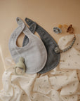 Two mushie Muslin Bibs, one in light gray and one in dark gray, are displayed on a white patterned cloth with a pacifier, pacifier holder, and silicone feeder, all set against a neutral beige background.