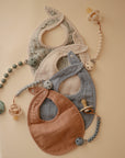 Three mushie Muslin Bibs in sage green, blue, and tan are arranged on a beige surface with matching beaded pacifier clips, while two pacifiers add a charming accent.
