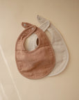 Two mushie Muslin Bibs made from soft, organic cotton with snap closures are shown overlapping on a beige background, in light brown and beige tones. Their minimalist design is perfect for teething babies.