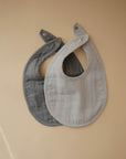 Two mushie Muslin Bibs, one gray and one light beige, are displayed on a beige surface. Made from organic cotton muslin, these bibs have snap closures and a simple, modern design.
