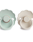 FRIGG Daisy Natural Rubber Pacifier 2-Pack by FRIGG features two daisy-shaped pacifiers—one light mint green and one cream—with scallop-edged shields, round handles, and natural rubber latex nipples, shown side by side on a white background.