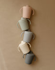 Five Mushie Trainer Sippy Cups in pastel pink, green, blue, cream, and gray with easy-grip handles are arranged vertically on a tan background. Each non-toxic cup is ideal for little hands.