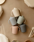 Five pastel Mushie Trainer Sippy Cups with easy-grip handles are arranged star-shaped on a beige surface, surrounded by matching Mushie plates, bowls, utensils, and containers in neutral tones.