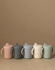 Five Mushie Trainer Sippy Cups with easy-grip handles in pastel pink, dark gray, light gray, cream, and sage green are arranged in a row against a beige background.