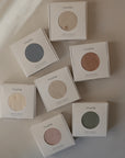 Seven boxes of mushie brand Stretchy Swaddle in various muted colors are arranged on a light surface, viewed from above. Each box has a round cutout showing the swaddle’s color, designed to provide a snug fit for your baby.